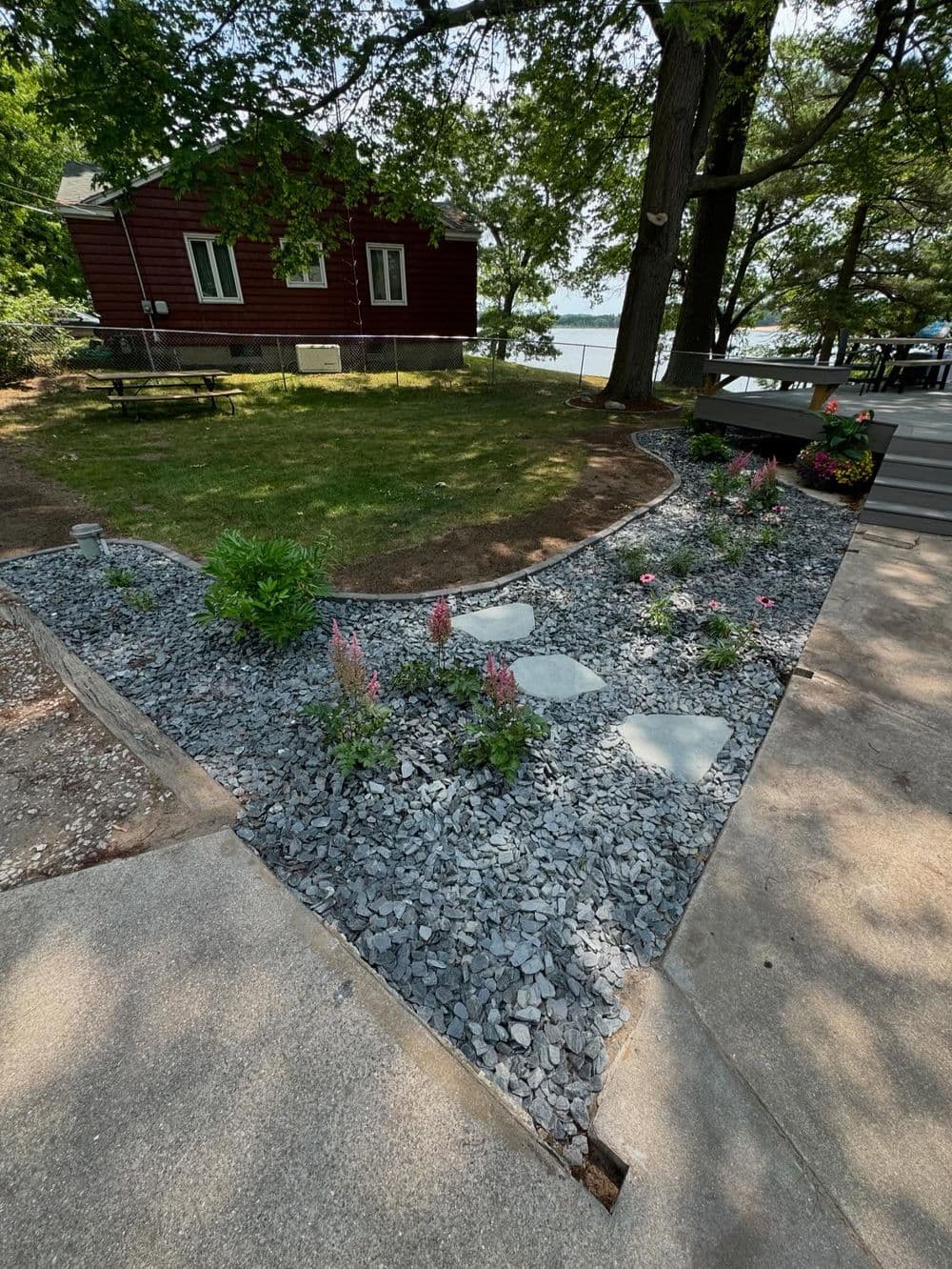Lush garden pathway with stone steps, bordered by colorful flowers and gravel, near a lakeside home.