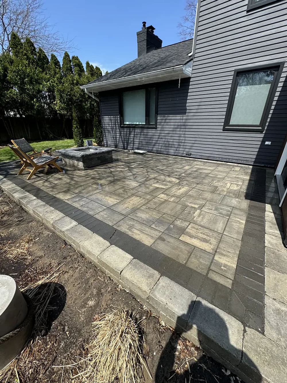 Outdoor patio with stone pavers, fire pit, and wooden chairs beside a modern home.