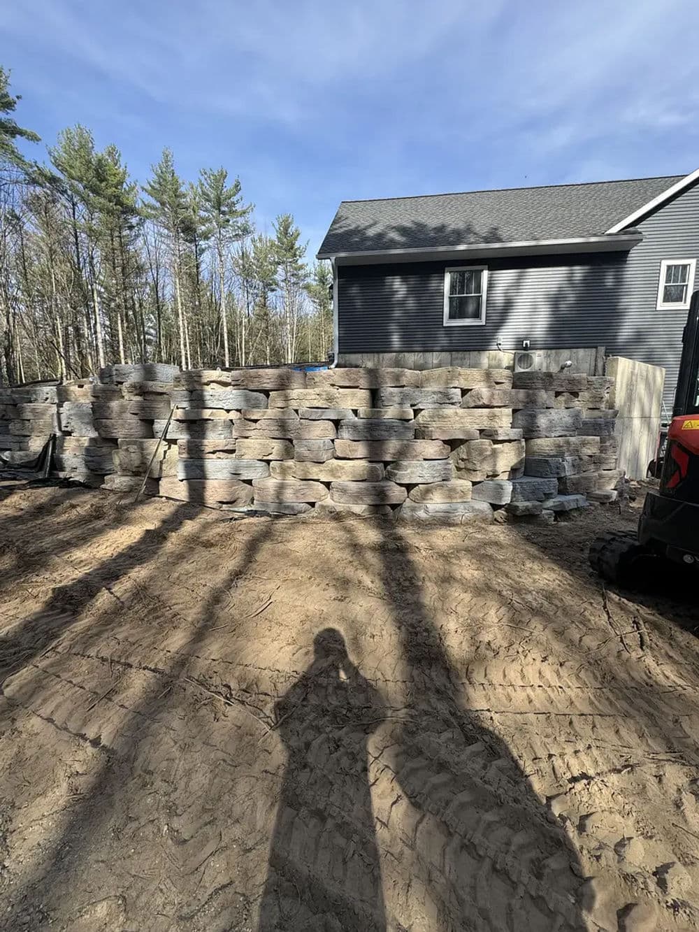 Stacked stone retaining wall behind a house surrounded by trees, with construction equipment nearby.