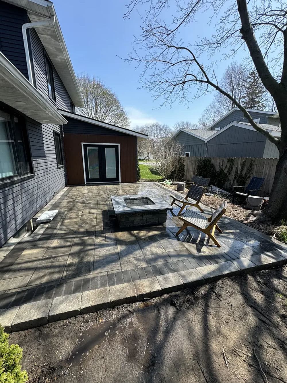 Patio area with fire pit, wooden chairs, and grassy landscape near a house on a sunny day.